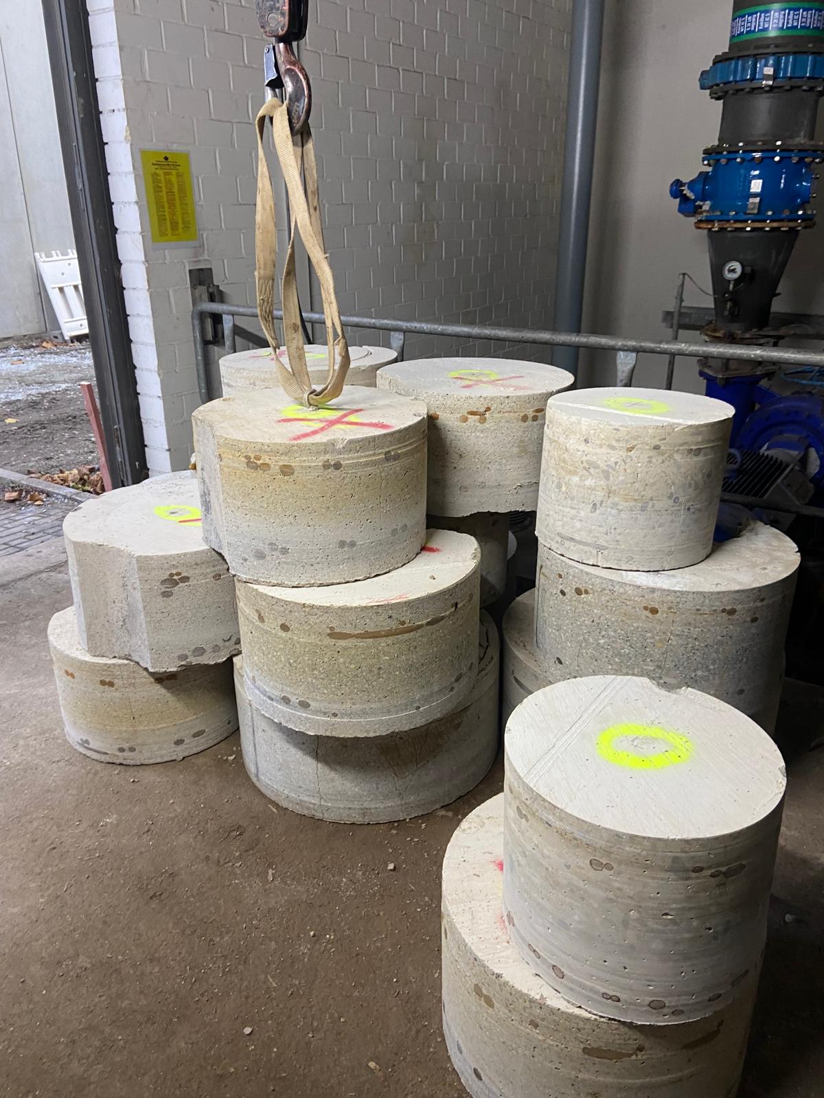ine Ansicht von mehreren großen, zylindrischen Betonbohrkernen, die in einem Industrieraum auf dem Boden gestapelt sind. Die Bohrkerne weisen Markierungen mit neongelber und roter Sprühfarbe auf. Ein Hebegurt hängt von einem Haken herab und ist um einen der oberen Betonblöcke geschlungen. Im Hintergrund sind eine weiß geflieste Wand, Metallgeländer sowie blaue Rohrleitungen und Flansche einer technischen Anlage erkennbar.