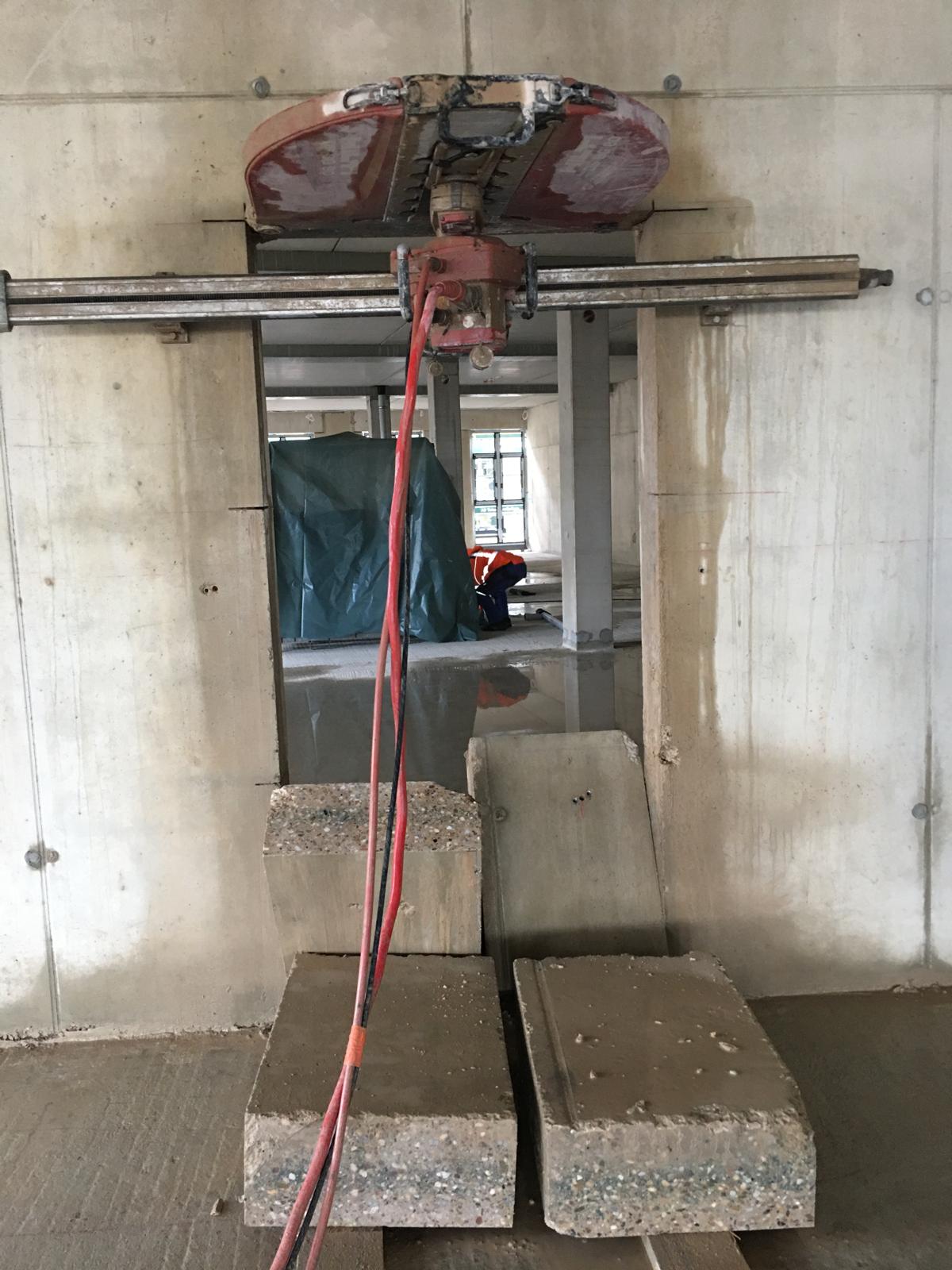 Eine professionelle Wandsäge mit einer großen, rötlichen Schutzhaube ist an einer Führungsschiene über einem bereits ausgeschnittenen Wanddurchbruch in einer Betonwand montiert. Zwei rote Versorgungsschläuche führen zum Sägekopf. Auf dem Boden vor der Öffnung liegen bereits herausgeschnittene, massive Betonblöcke. Durch den Durchbruch ist im Hintergrund eine weitere Baustellenszene mit Pfeilern und einer grünen Abdeckplane erkennbar.