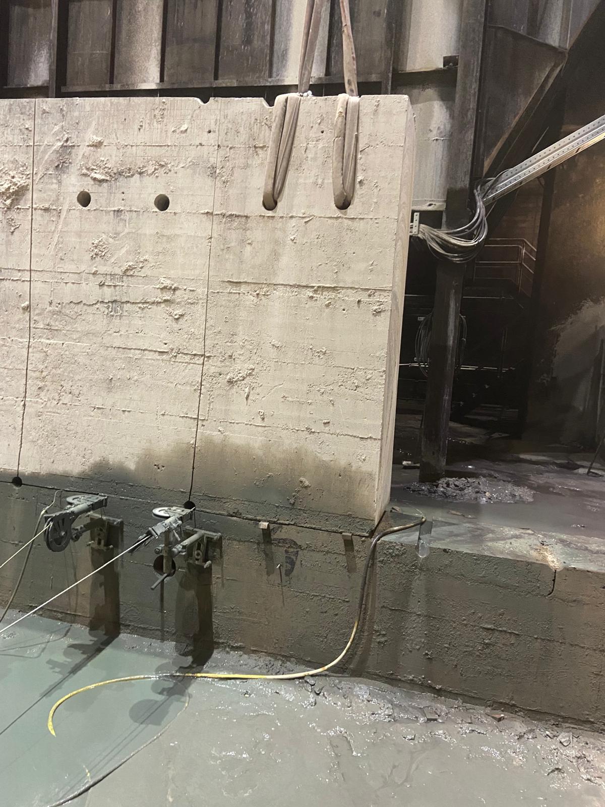 Eine Detailaufnahme einer Betonwandsäge im Einsatz bei einer industriellen Demontage. Ein massiver Betonblock wird mithilfe von zwei breiten Hebegurten, die durch Kernbohrungen im oberen Bereich gefädelt sind, gesichert. Im unteren Bereich der Wand sind die Schienen und Führungsköpfe einer Diamant-Seilsäge oder Wandsäge montiert, die einen präzisen vertikalen Schnitt ausführen. Am Boden hat sich grauer Betonschlamm angesammelt, und im Hintergrund ist eine dunkle Stahlkonstruktion erkennbar.