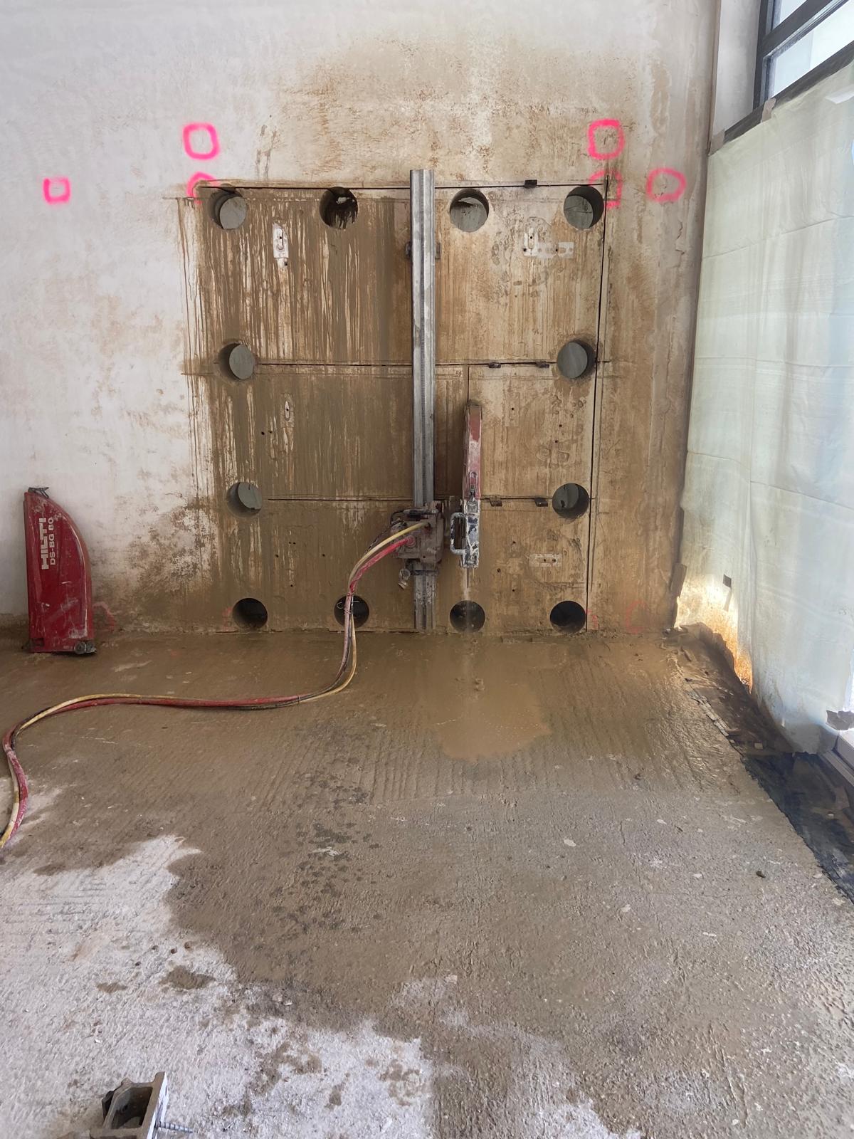 Eine Innenaufnahme einer Baustelle während eines Wanddurchbruchs. Eine vertikale Führungsschiene einer Betonsäge ist mittig auf einer markierten Wandfläche montiert. Die Wand ist ringsherum mit präzisen Kernbohrungen versehen und weist deutliche Schlammspuren vom wassergekühlten Sägeprozess auf. Links steht eine rote Schutzhaube für das Sägeblatt am Boden, und pinkfarbene Sprühmarkierungen kennzeichnen den Arbeitsbereich. Der Boden davor ist mit einer dünnen Schicht aus Betonschlamm bedeckt.