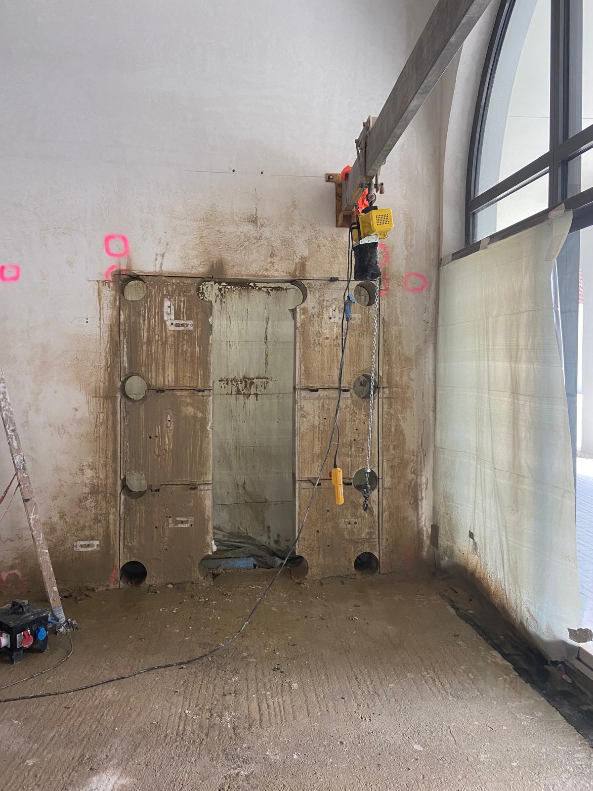 Eine Innenaufnahme einer Baustelle, die eine Wand mit einer vorbereiteten Wandöffnung zeigt. Um die Öffnung herum wurden präzise Kernbohrungen vorgenommen, und die Wandfläche weist Schlammspuren vom wassergekühlten Schneidprozess sowie pinkfarbene Markierungen auf. Über der Öffnung ist ein gelber Elektrokettenzug an einem Metallausleger montiert, der zur Lastenhebung dient. Ein großes Fenster auf der rechten Seite ist teilweise mit einer hellen Plane abgehängt, und auf dem Boden liegen Kabel sowie ein Baustromverteiler.