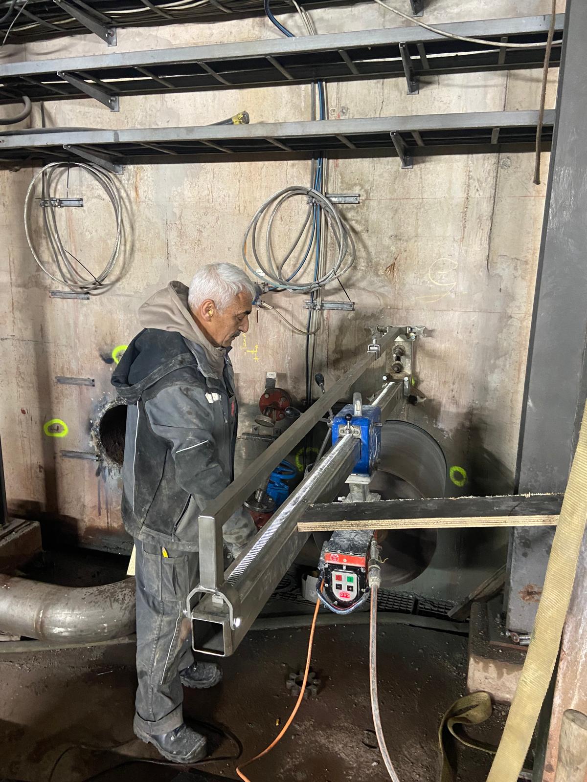 Ein erfahrener Arbeiter bedient eine Kernbohrmaschine, die horizontal an einer massiven Betonwand in einem Technikraum montiert ist. Die Maschine führt gerade eine Bohrung mit großem Durchmesser durch. Der Fachmann trägt graue Arbeitskleidung und Gehörschutz. Im Hintergrund sind bereits eine fertige Kernbohrung, verschiedene Kabelstränge an der Wand sowie Rohrleitungen im unteren Bereich erkennbar. Der Arbeitsbereich wird durch künstliches Licht beleuchtet.