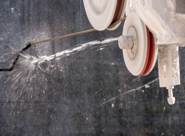 Eine Nahaufnahme einer Diamantseilsäge im Einsatz, die gerade einen massiven, dunklen Steinblock schneidet. Das Seil wird über zwei große, helle Umlenkrollen mit roten Gummieinlagen geführt. An der Stelle, an der das Seil in den Stein eintritt, wird Wasser zur Kühlung und Staubbindung aufgespritzt, das effektvoll zur Seite spritzt. Die Szene verdeutlicht die Präzision und den technischen Aufwand beim Trennen von extrem harten Materialien in Steinbrüchen oder bei Spezialabbruch-Projekten.